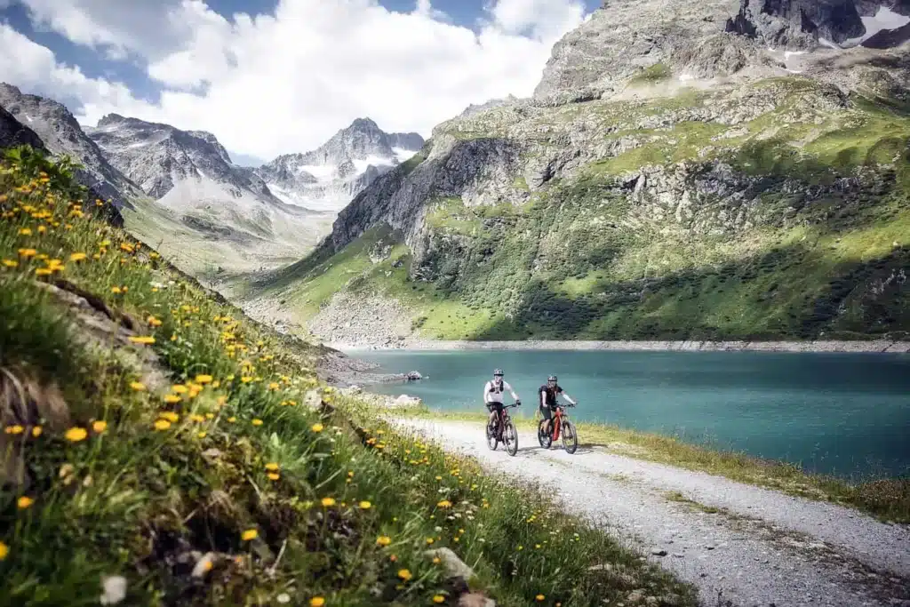 Two mountain bikers riding a lakeside gravel path beside a turquoise alpine lake with wildflowers and dramatic rocky peaks in St. Anton.