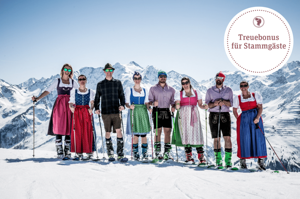 Group in traditional dirndl and lederhosen with ski poles posing on a snowy mountain, promoting a 'Treuebonus für Stammgäste' loyalty bonus offer.