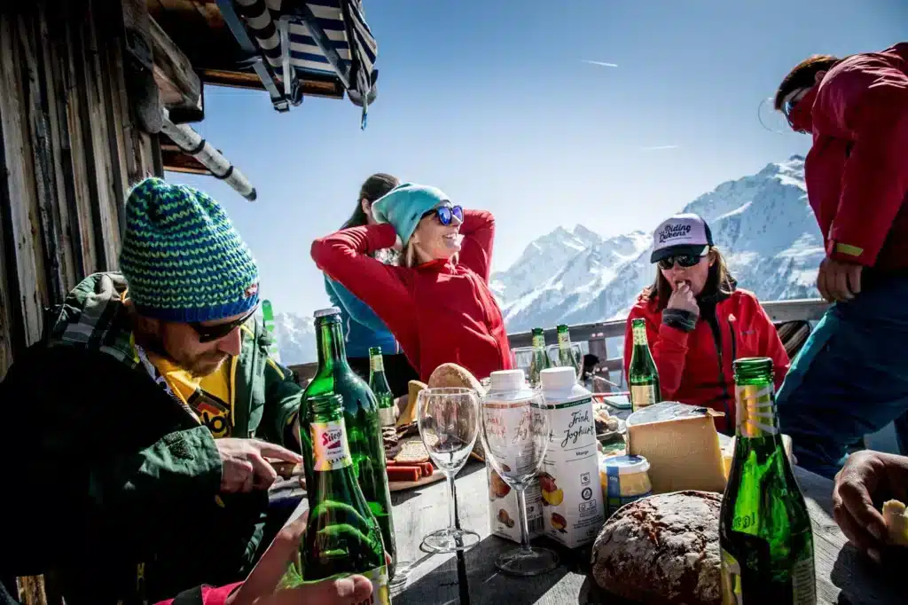 Skiers relaxing at mountain hut table with bread, cheese, wine bottles, snowy peaks backdrop.
