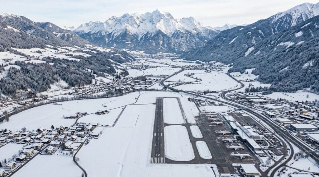 Aerial winter view of Innsbruck Airport with snow-covered runway, parked planes, surrounding villages, and dramatic snow-capped alpine mountains in the background.