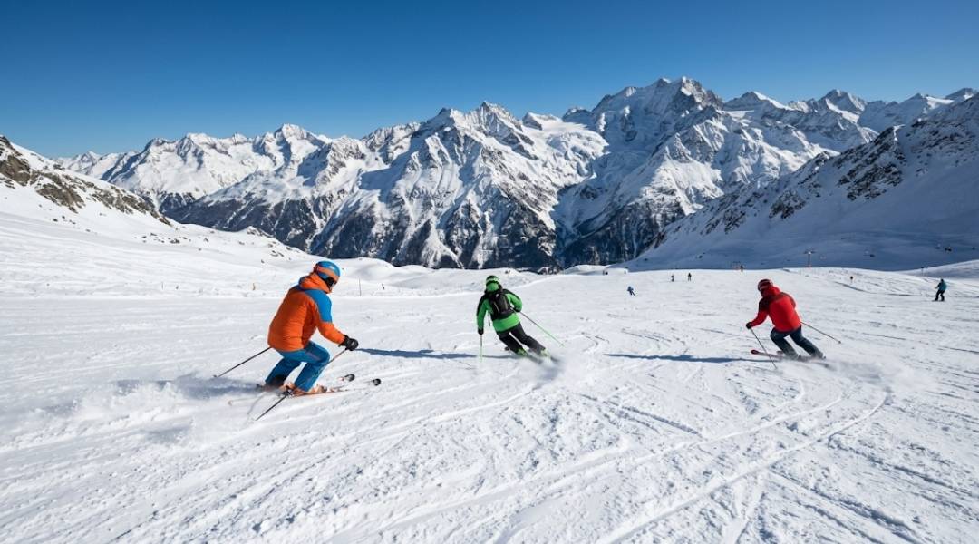 Three skiers carving fresh snow on a sunny St. Anton am Arlberg piste with dramatic Alpine peaks — perfect conditions for the best time to ski.
