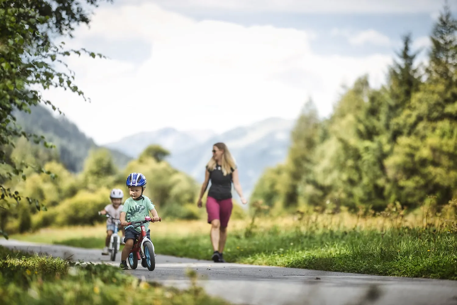 Homepage 17 Woman walking behind two young children in helmets riding balance bikes on a paved path near mountains