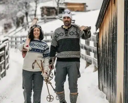 Homepage 15 Smiling couple in patterned wool sweaters carrying vintage wooden skis and poles along a snowy lane beside a barn